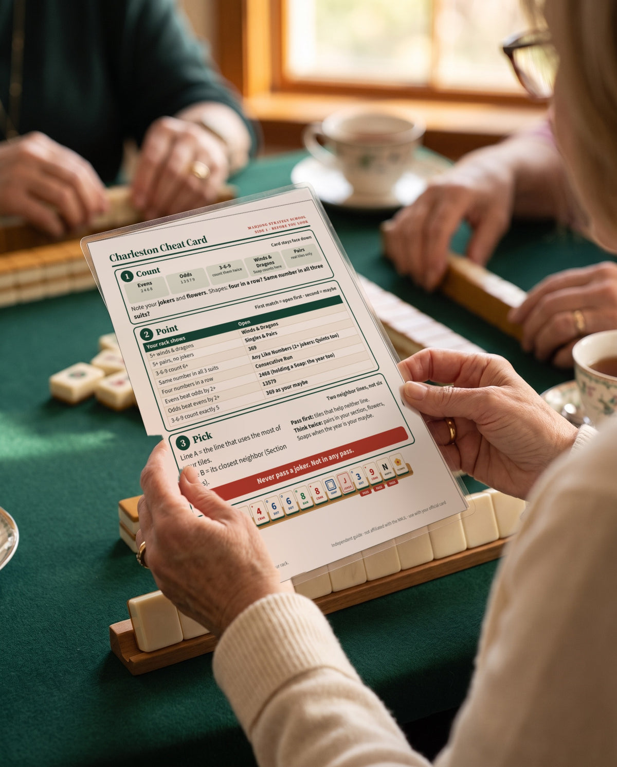 A player holds the laminated Charleston Cheat Card over her rack at a mahj table